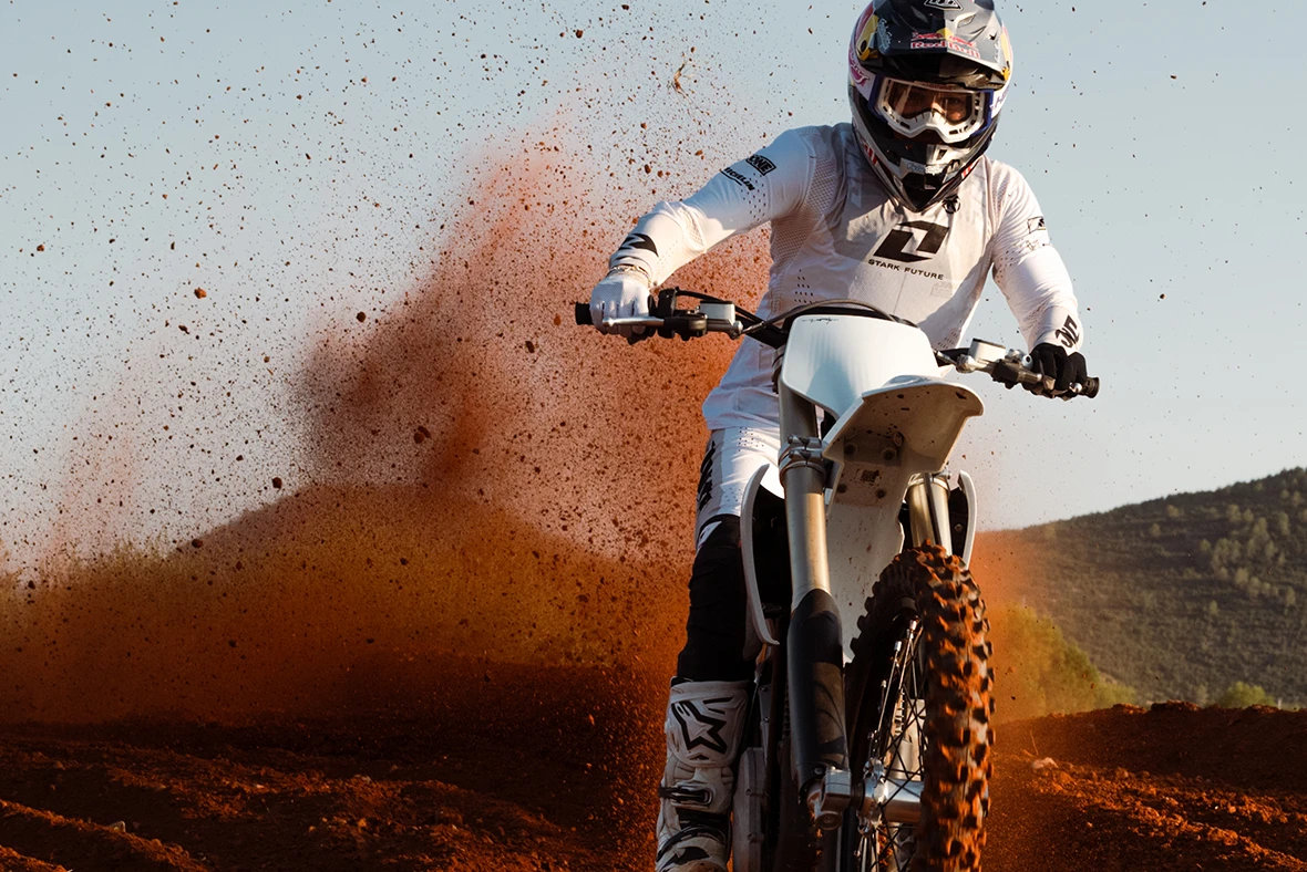A rider in white gear standing on a VARG and kicking up red dust on a dirt track.